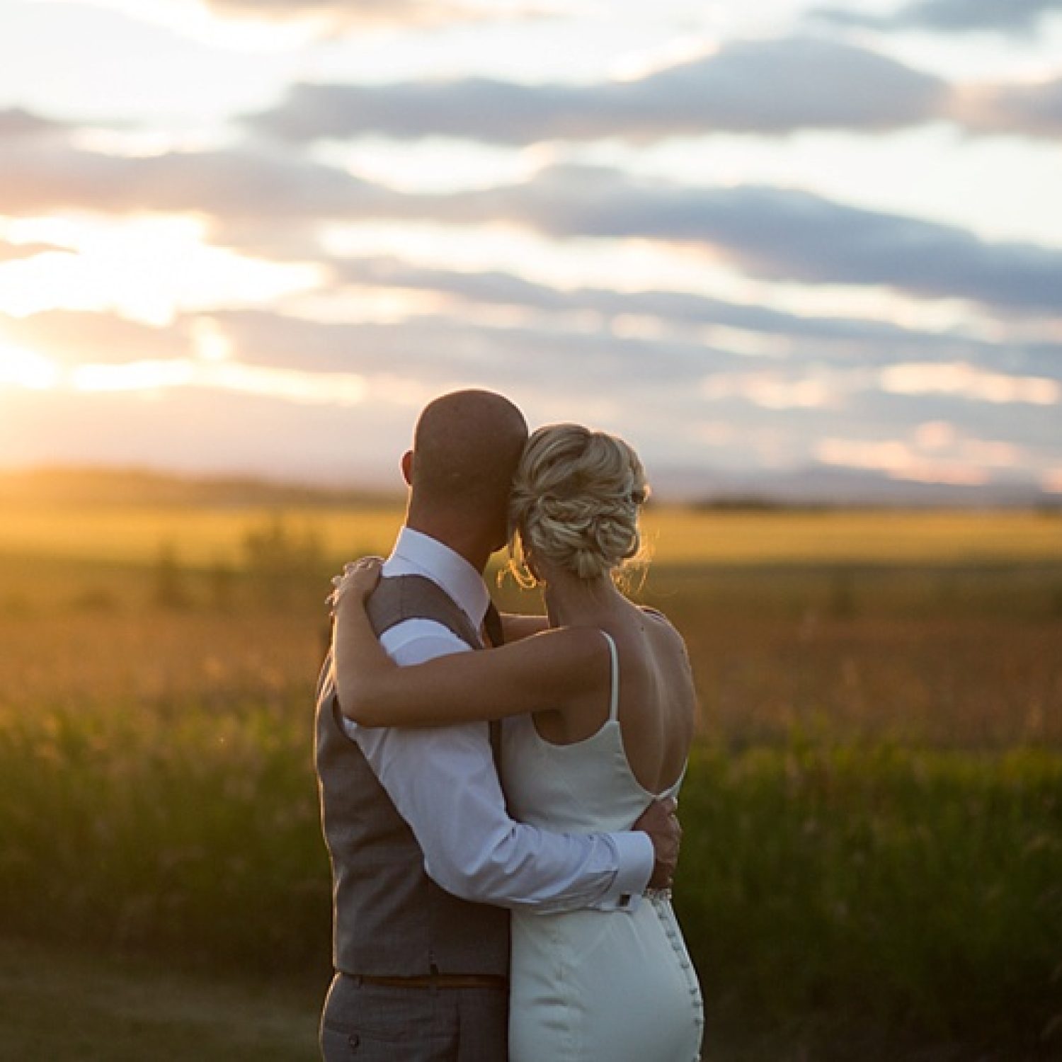 Romantic golden hour wedding photo of a couple embracing in a field at sunset, highlighting the soft natural light at Bowness Park.